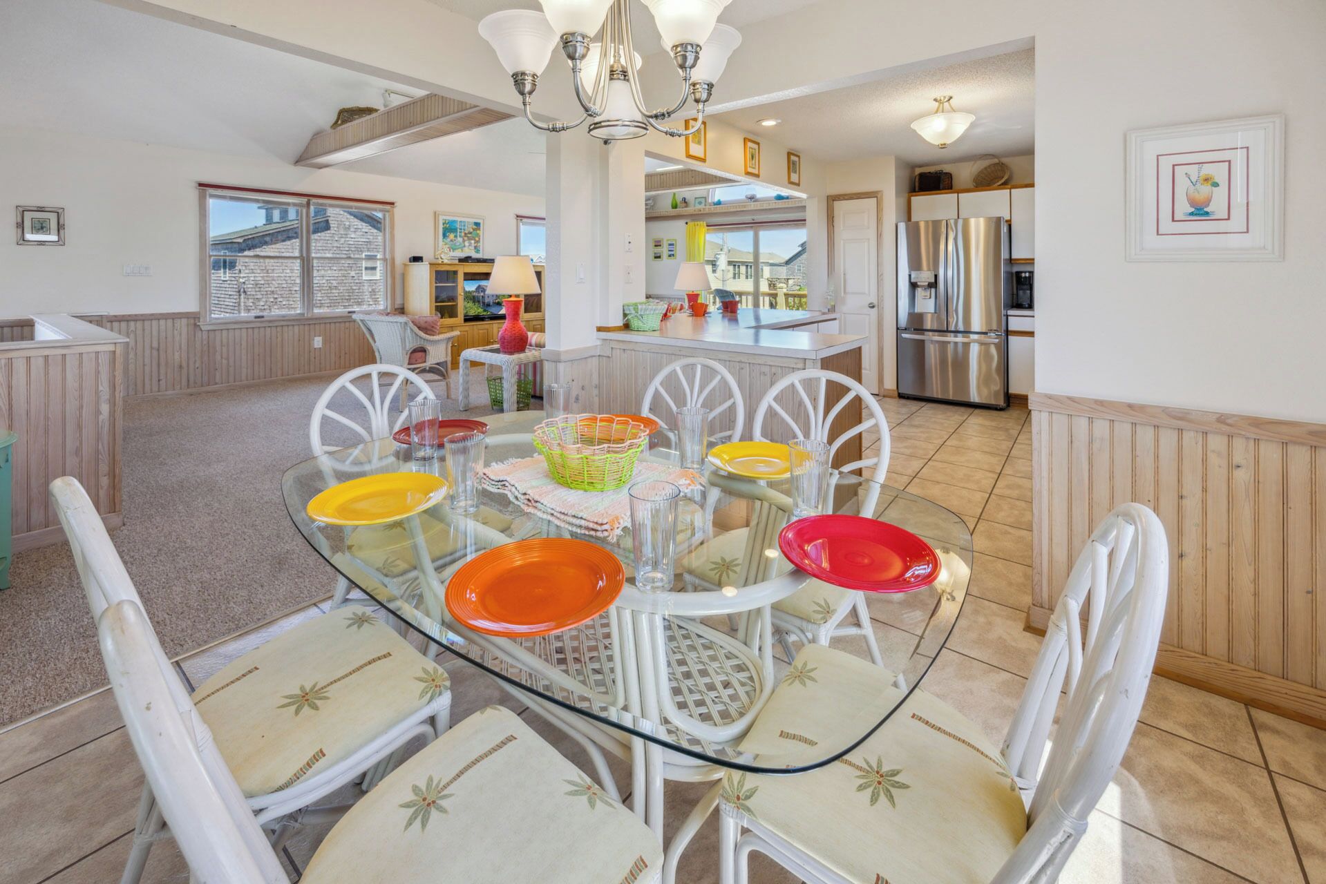 A dining area with a glass table and white chairs, a kitchen in the background, and a view of a house through a window.