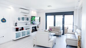 A modern living room with a sofa, armchairs, a coffee table, and a TV. There is a large window with a view of the outdoors. - Luxury Apt in the center of Glyfada (Glyfada)
