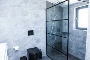 A modern bathroom with grey tiles, a glass-enclosed shower, a black toilet, and a window providing natural light. - Luxury Apt in the center of Glyfada (Glyfada)