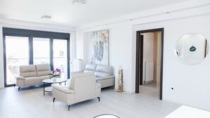 A modern living room with a sofa set, a coffee table, and a floor lamp. There is a large window providing natural light and a view of the outside. - Luxury Apt in the center of Glyfada (Glyfada)
