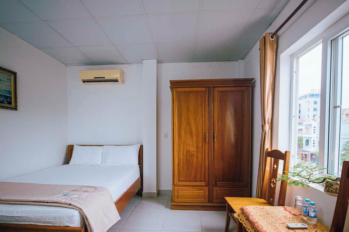 Un dormitorio con cama, armario, mesa con planta y ventana con vistas a edificios.