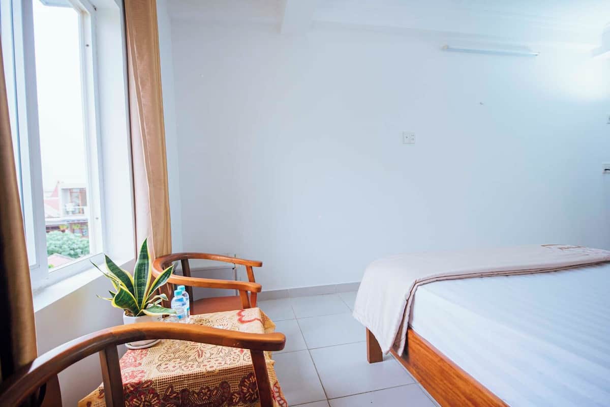 Habitación de hotel con cama, una silla, una mesita con planta y botellas de agua, y una ventana con cortinas.