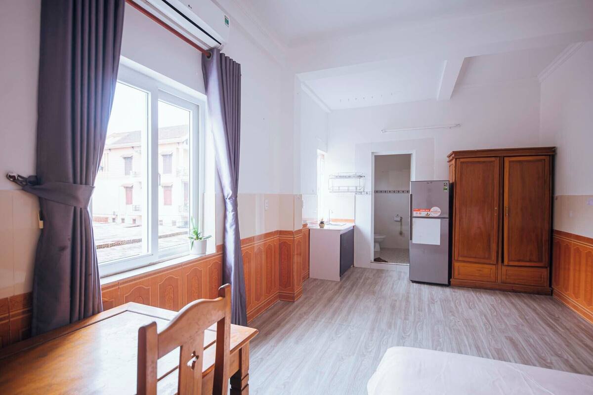 Habitación de hotel con una mesa de comedor de madera, una cama, un armario, una pequeña kitchenette y una ventana con cortinas.