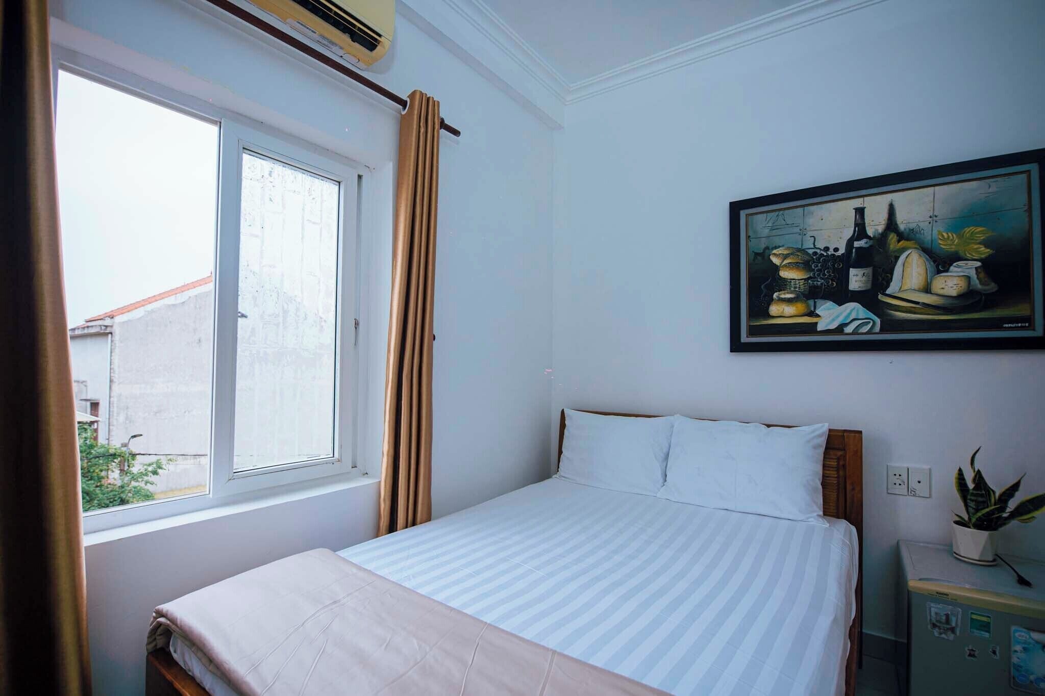 A hotel room with a bed, a window with curtains, a painting on the wall, and a small table with a plant.