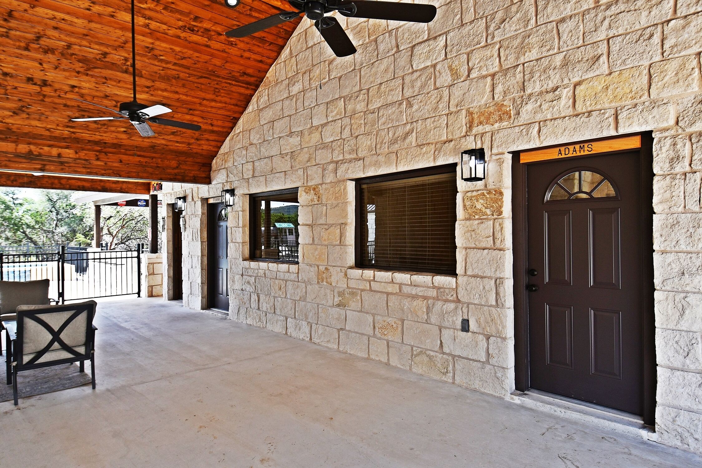 A stone building with a dark brown door labeled "Adams," a covered patio area with a ceiling fan, and a window with blinds.