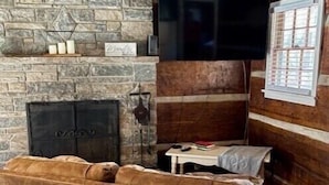 A cozy living room with a stone fireplace, a flat-screen TV mounted above it, and a brown leather sofa. - Cabin 5 - Rustic cabin in Pioneer Village, Great Smoky Mountains NC (Maggie Valley)