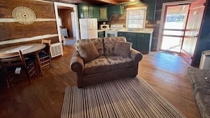 A cozy living room with a sofa, a dining table, and a kitchen area with cabinets and a refrigerator. - Cabin 5 - Rustic cabin in Pioneer Village, Great Smoky Mountains NC (Maggie Valley)