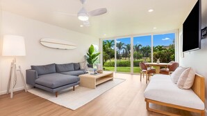 A modern living room with a grey sofa, a wooden coffee table, and a white sofa. There is a ceiling fan, a TV mounted on the wall, and a large window with a view of palm trees. - Hale Maluhia ~ Peaceful home (Kawela Bay)