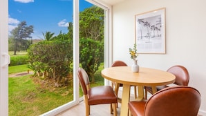 A dining area with a round table and six brown chairs, a framed picture on the wall, and a view of a garden through a large window. - Hale Maluhia ~ Peaceful home (Kawela Bay)