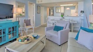 Ein modernes Wohnzimmer mit Sofa, Sessel und Couchtisch. Im Hintergrund befindet sich ein Essbereich und ein Fernseher auf einem Schrank.