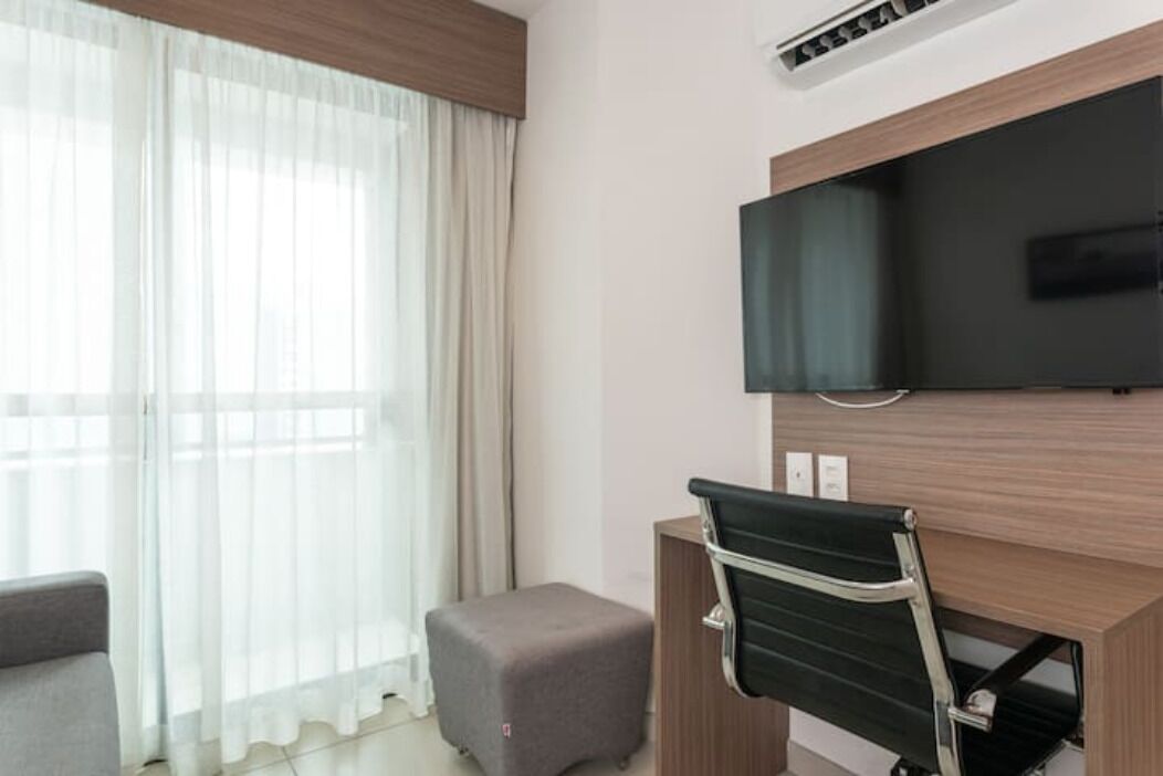 Habitación de hotel con cama, televisor montado en la pared, escritorio con silla y ventana con cortinas traslúcidas.