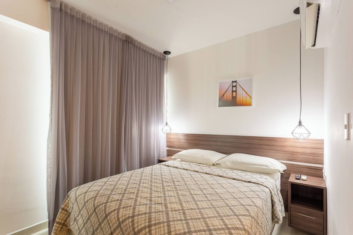 A neatly arranged bedroom with a large bed, a nightstand, a wall-mounted air conditioner, and a painting of a bridge.