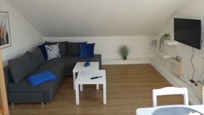 Ein modernes Wohnzimmer mit einem grauen Ecksofa, einem weißen Couchtisch und einem weißen Sessel. Ein Flachbildfernseher ist an der Wand befestigt und auf einem Regal steht eine kleine Pflanze.