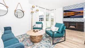 Een moderne woonkamer met blauwgroene banken, een ronde houten salontafel en een groot televisiescherm waarop een zonsondergang te zien is.