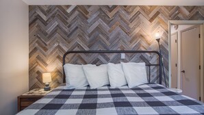 Una cama con colcha a cuadros, una mesita de noche con lámpara y una pared de madera con motivo de espiga.