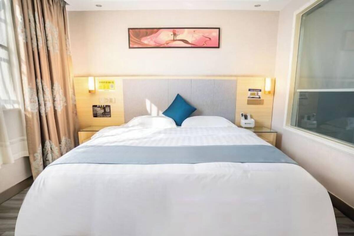 A hotel room with a large bed, a headboard, bedside tables, a nightstand, a window, and a painting on the wall.