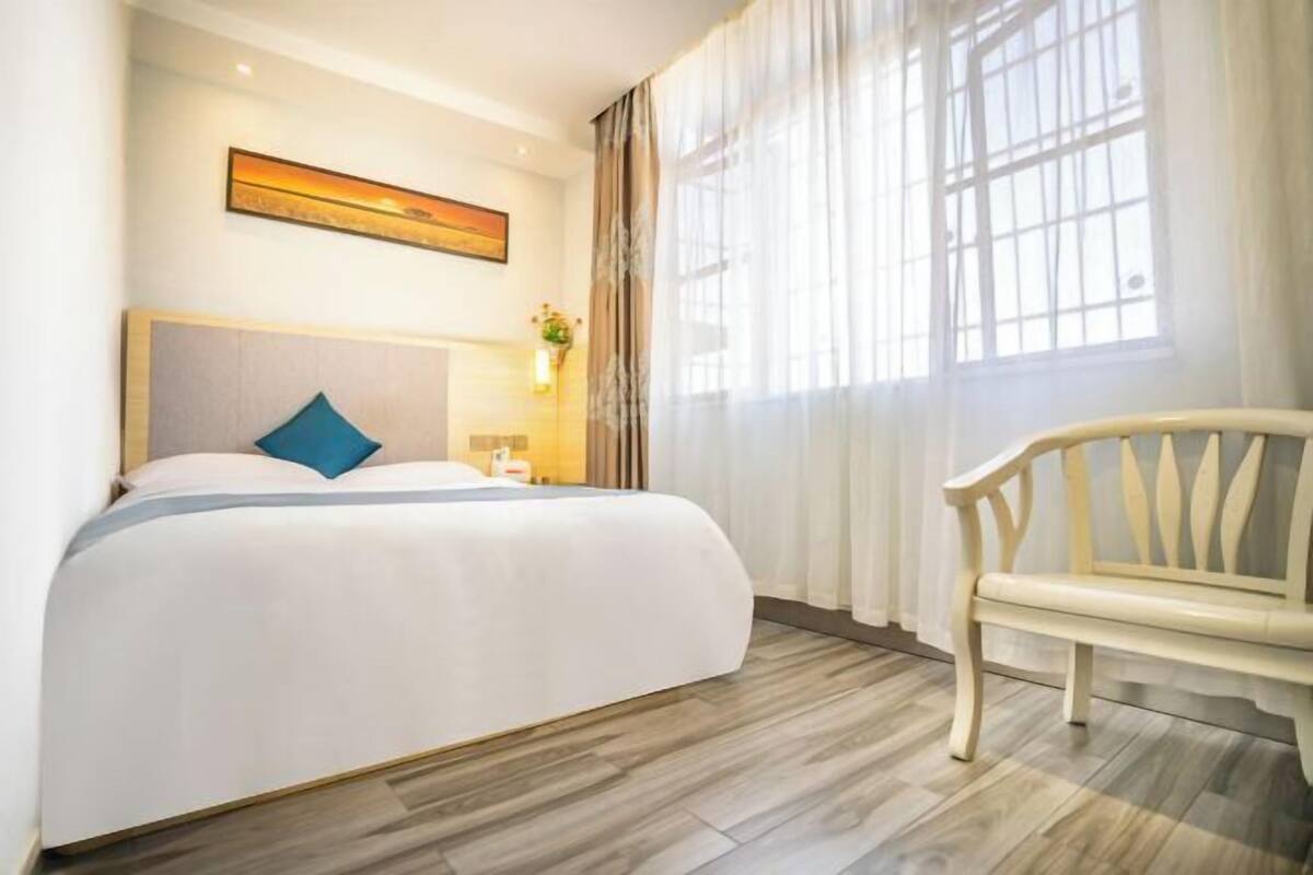 A hotel room with a bed, a bench, a painting on the wall, and a window with curtains.