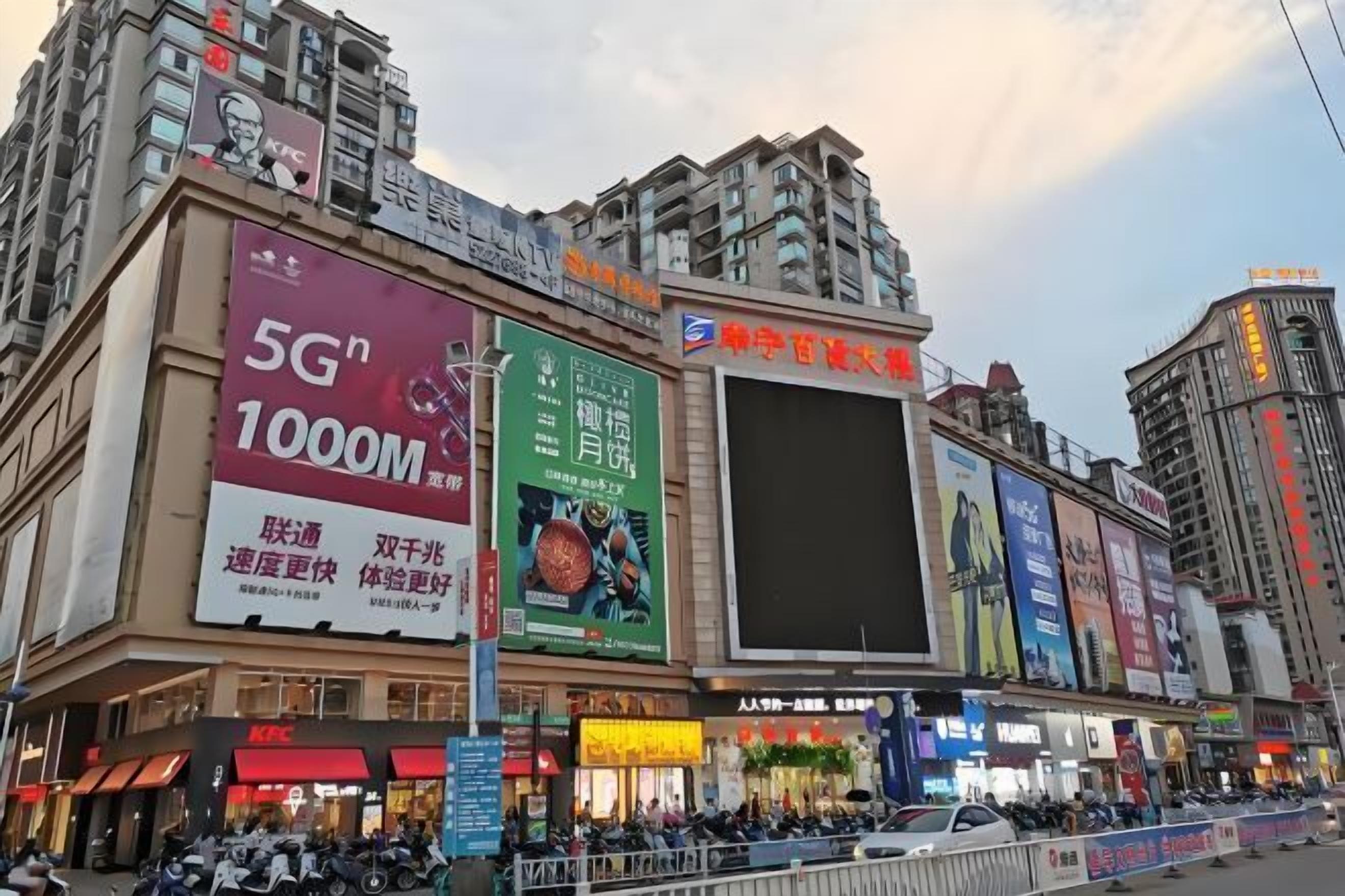 A busy urban street with multiple commercial buildings, including a prominent KFC restaurant and a large digital billboard advertising 5G technology.