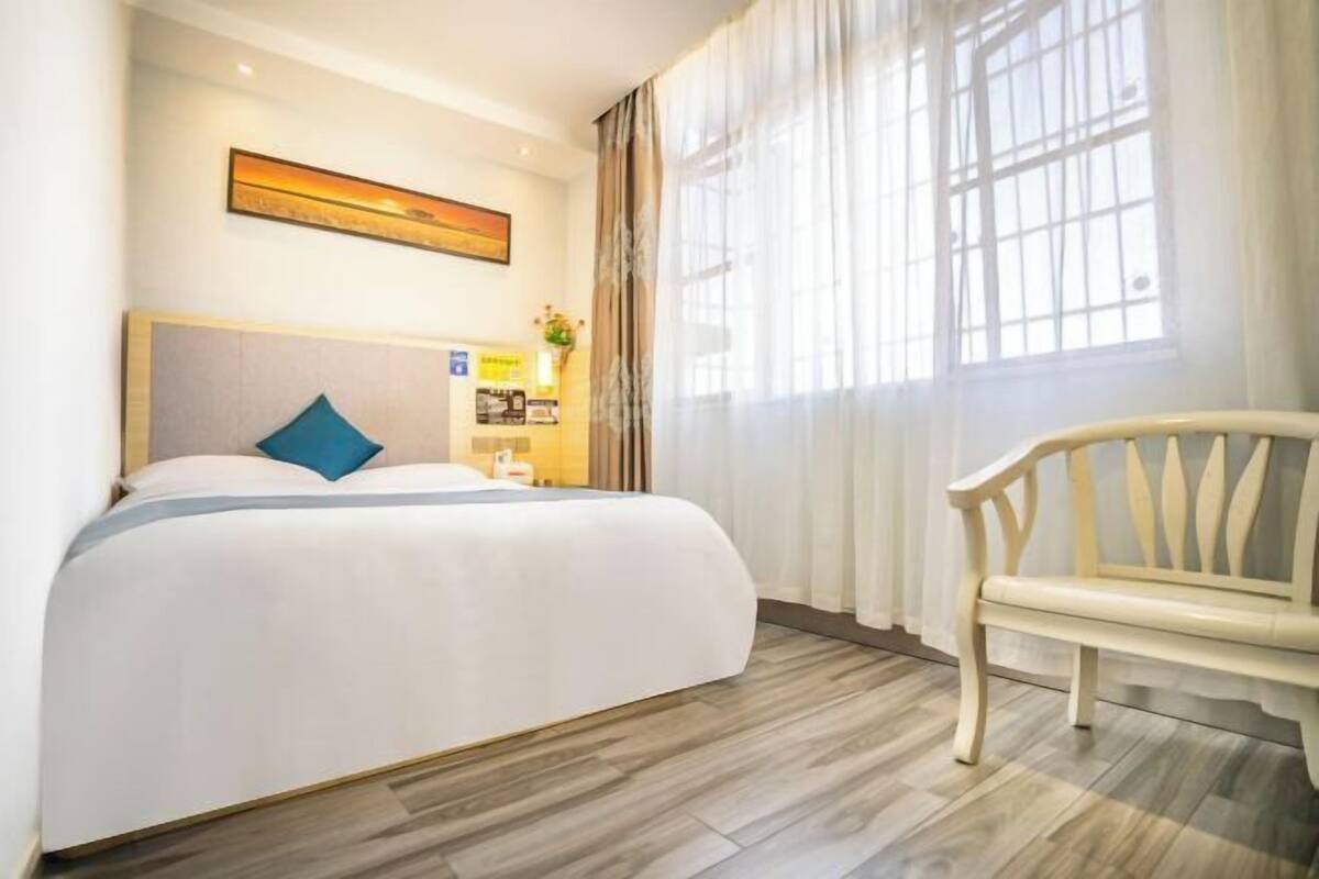 A hotel room with a large bed, a wooden bench, a desk with a lamp, and a window with curtains.