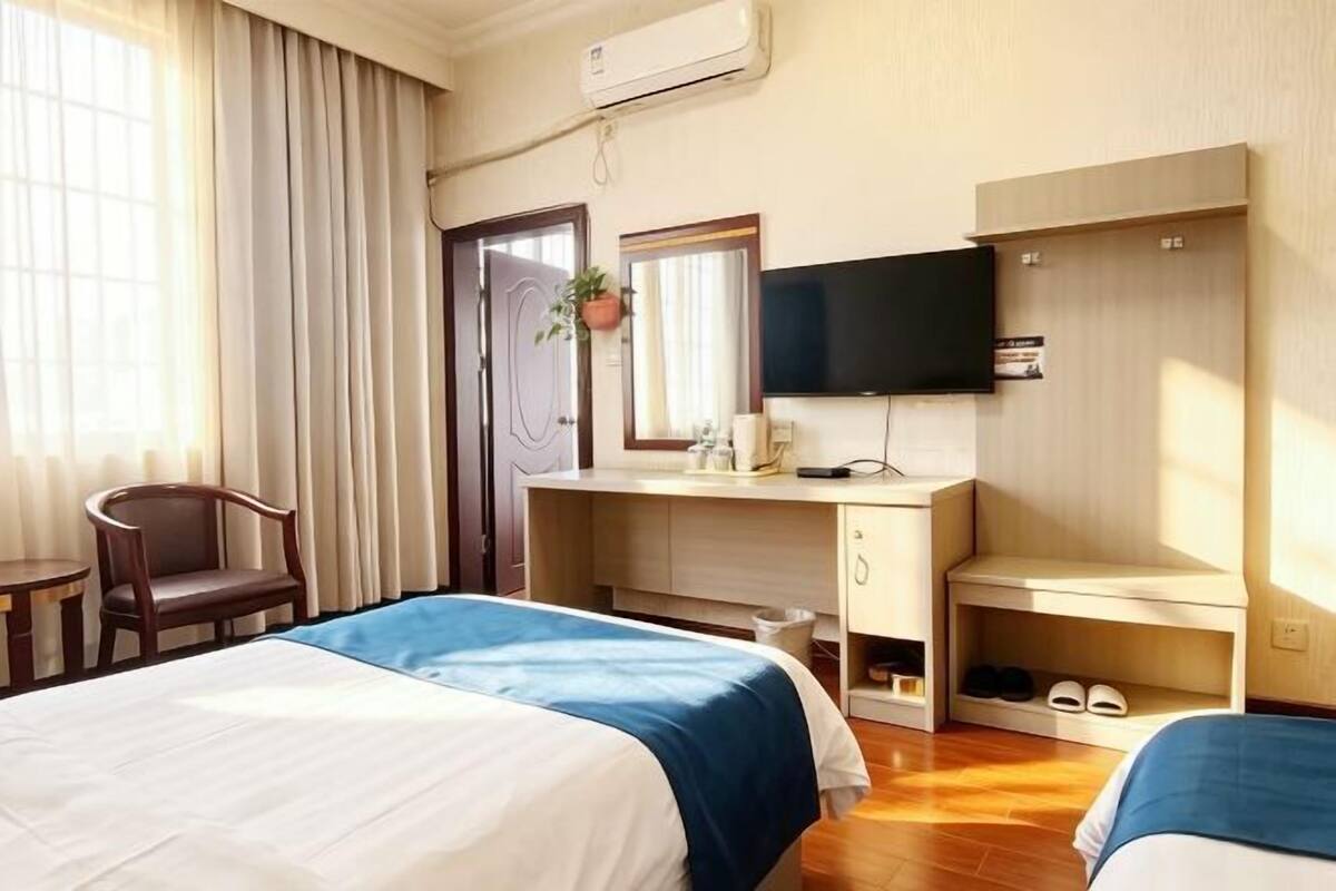A hotel room with two beds, a desk, a TV, a mirror, a plant, and a shoe rack.