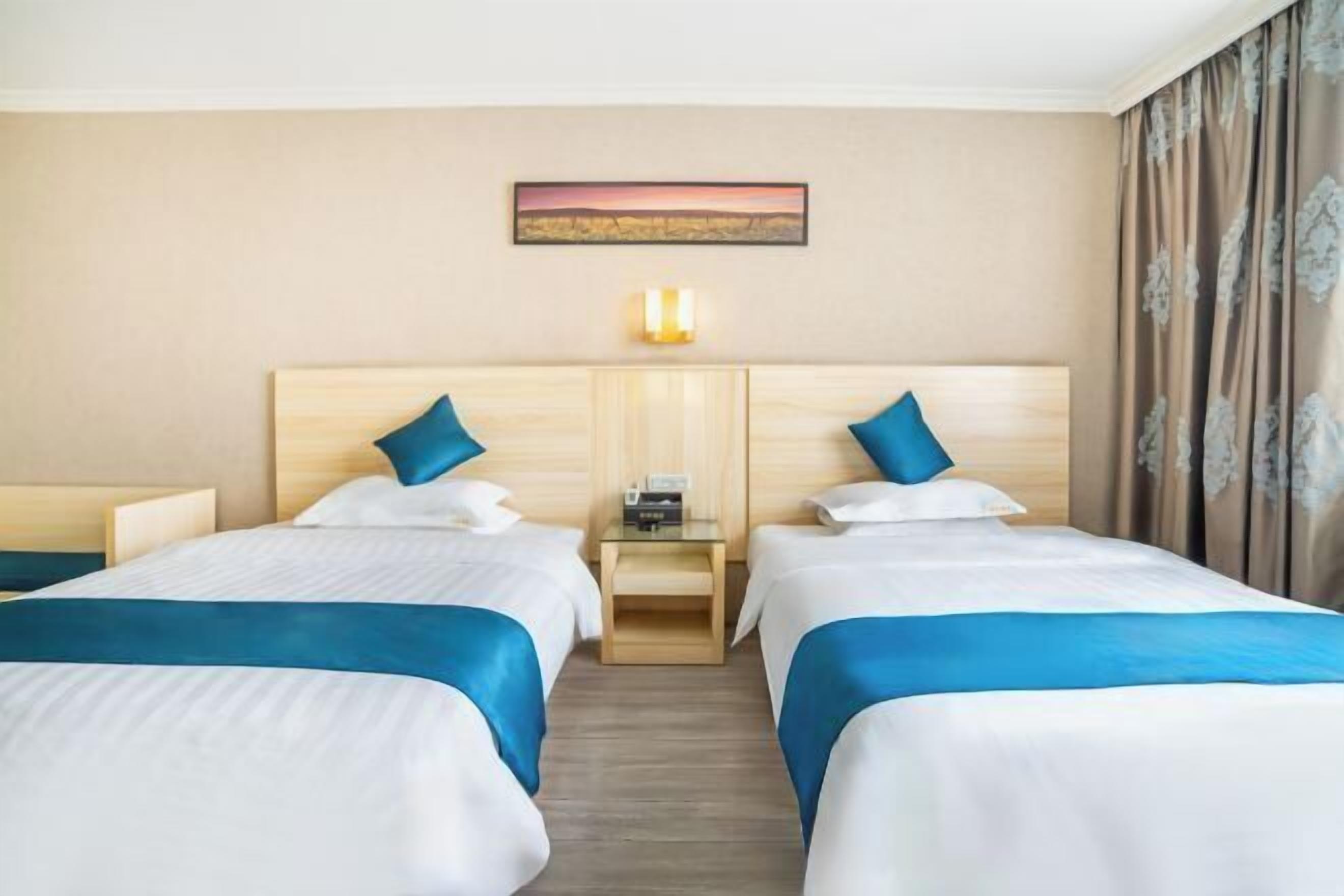 A hotel room with two beds, each with blue and white bedding, a wooden headboard, a bedside table with a lamp, and a painting on the wall.