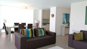 A modern living room with a brown sofa, a dining table with chairs, and a kitchen area in the background. - Seaview and private pool - house for 6 (Lourinhã)