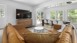 Una sala de estar moderna con un televisor de pantalla plana grande, un sofá de cuero marrón, una mesa de centro redonda y un comedor con una mesa y sillas de madera.