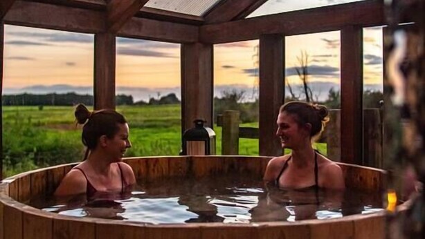 A wooden hot tub with a view of a sunset over a green landscape.
