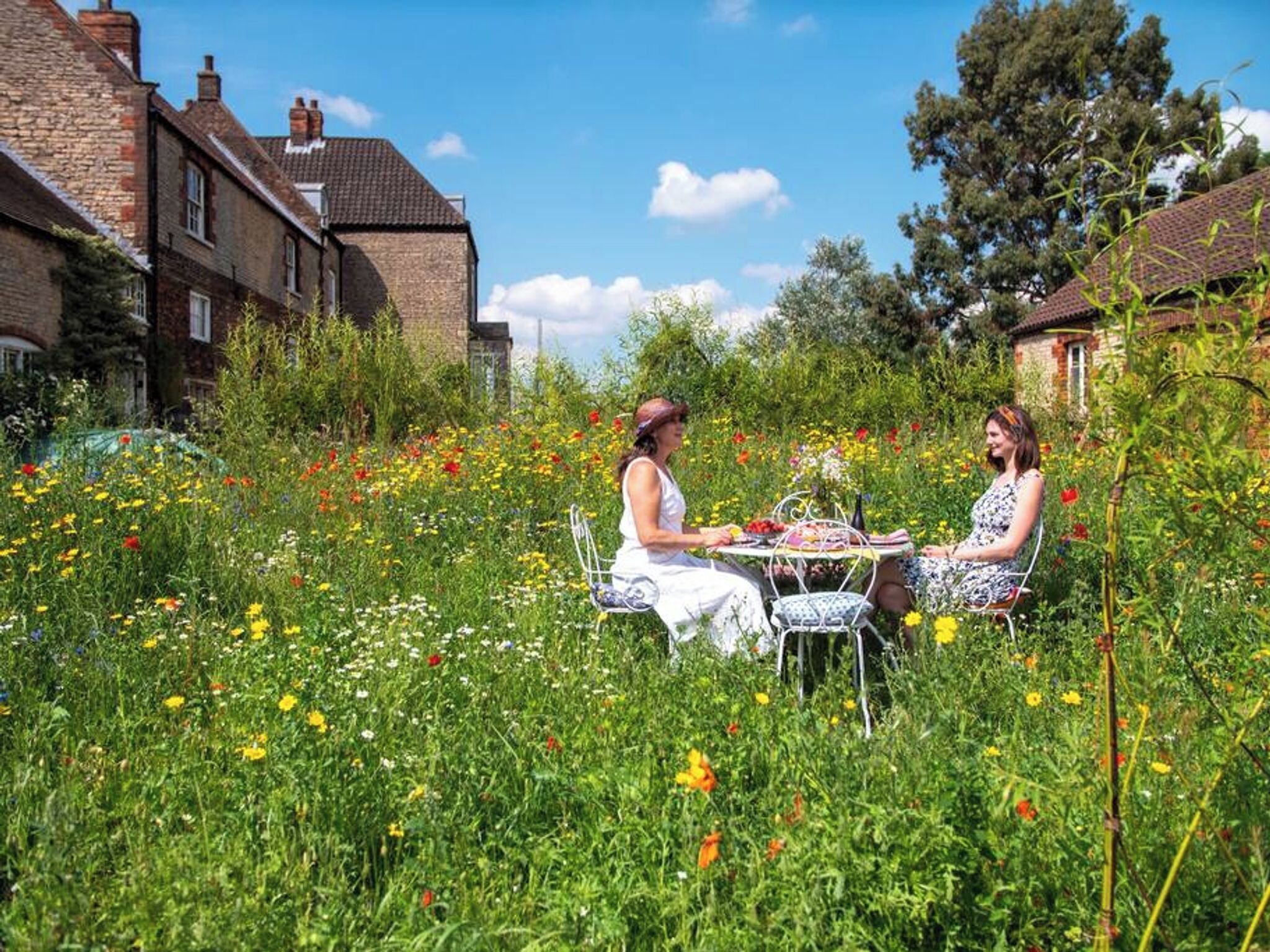 Zwei Personen sitzen an einem Tisch in einem Garten voller Blumen, im Hintergrund ein Backsteinhaus.