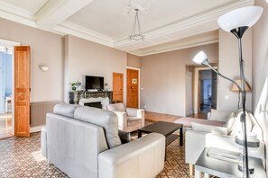 Amplia sala de estar con sofá, sillones y una mesa de centro. Un televisor está montado en la pared y hay una araña sobre la zona de estar.