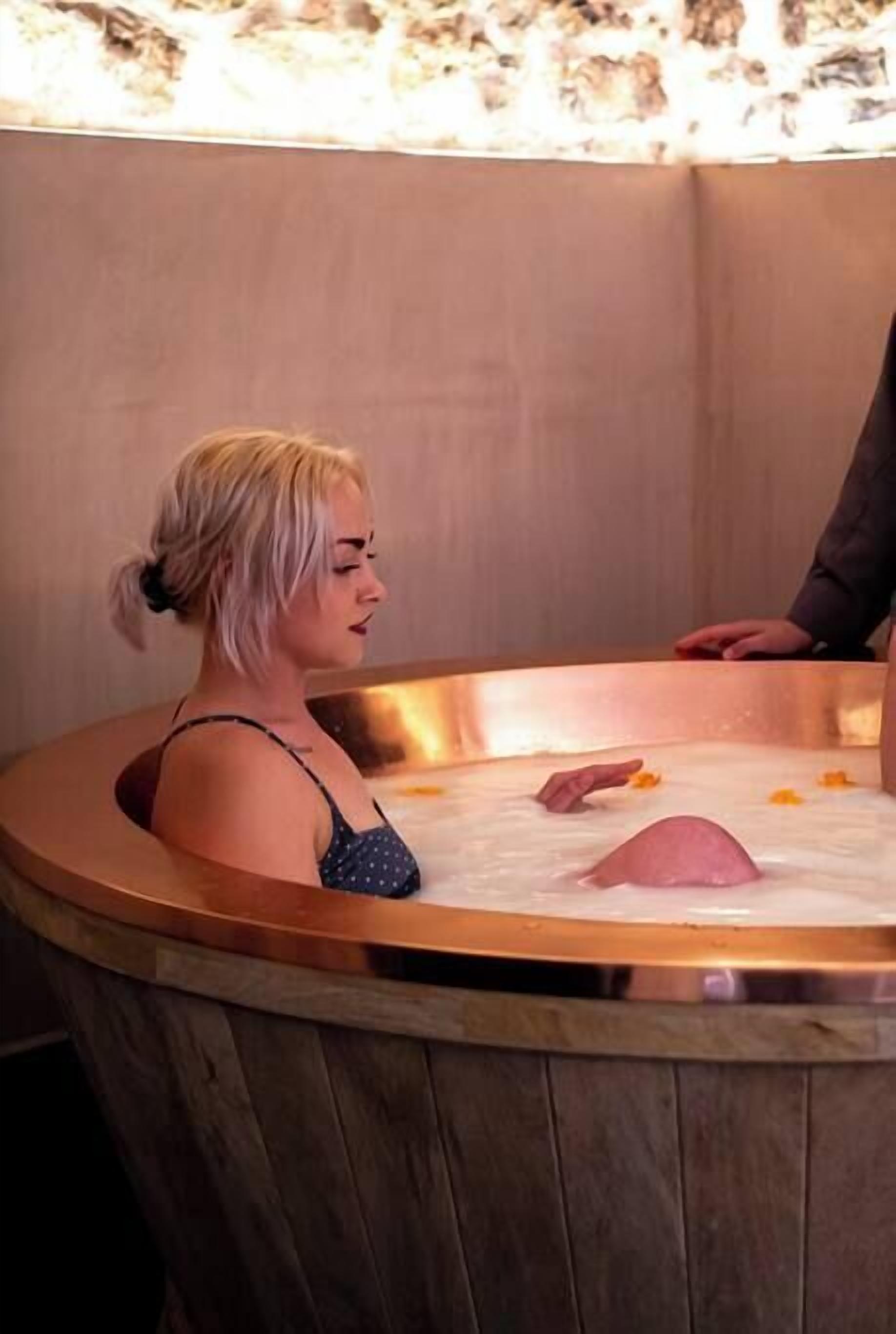 Una donna in una vasca da bagno di legno con una persona accanto.