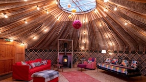 Un intérieur chaleureux d’une yourte, avec un foyer central, des murs en bois et un canapé rouge.