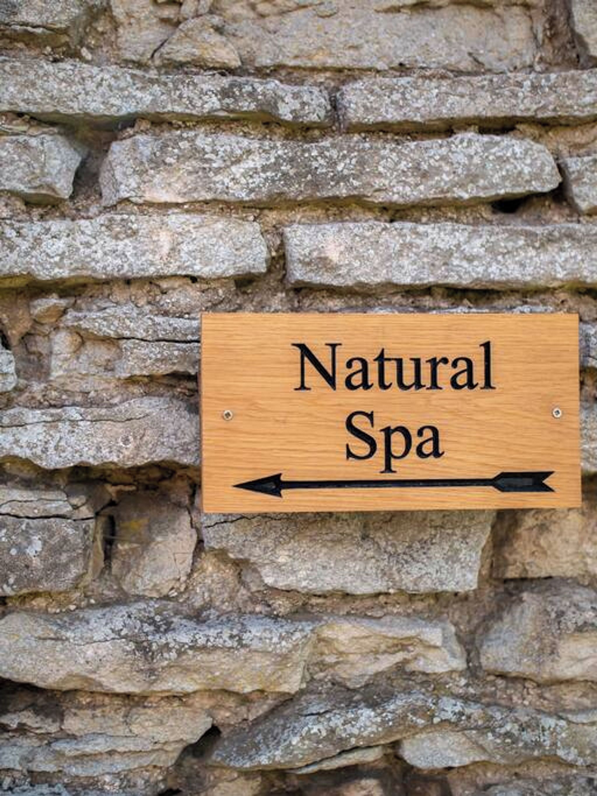 Uma placa de madeira com o texto 'Natural Spa' e setas indicativas, ao fundo de uma parede de pedra.