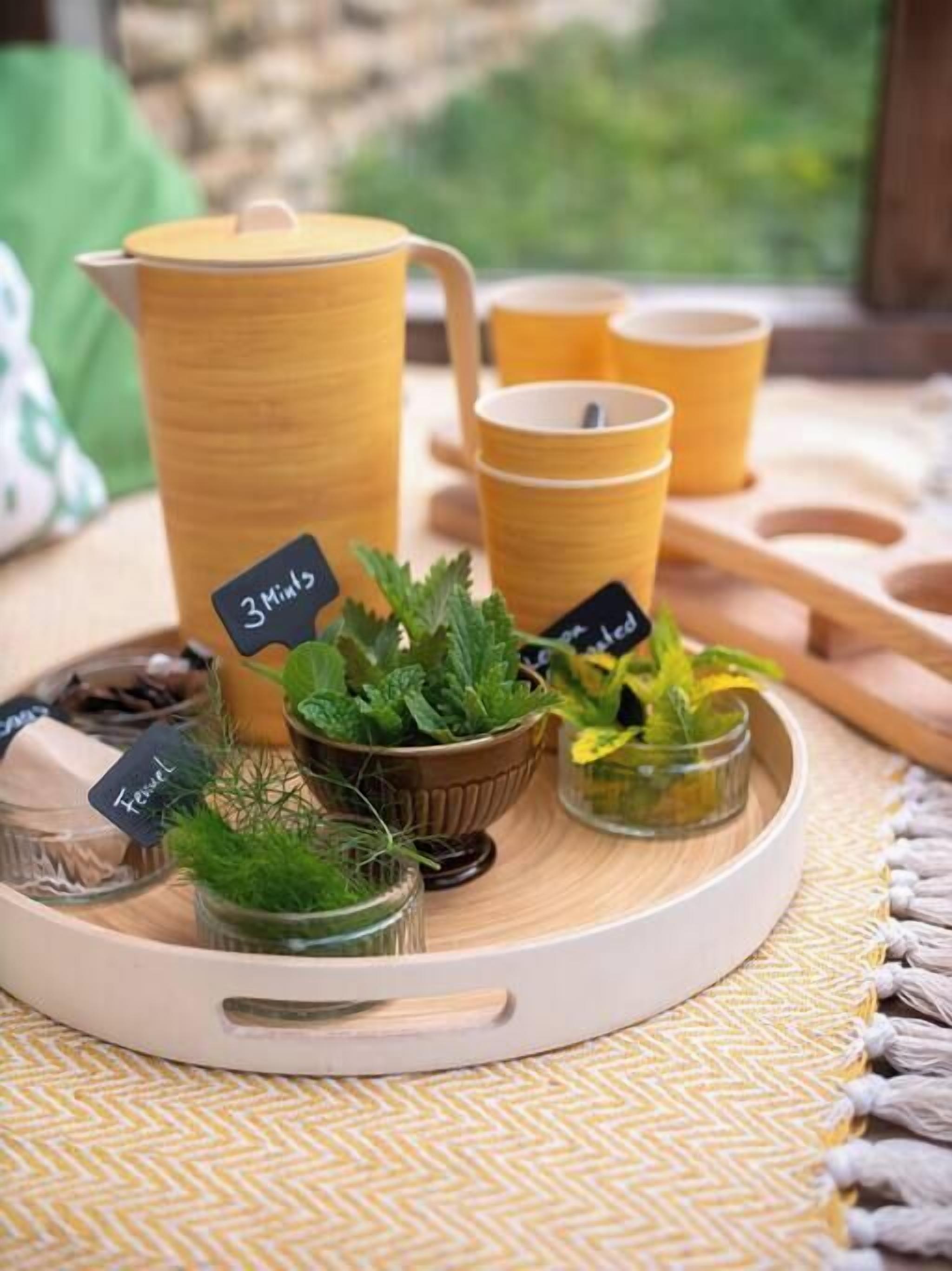 Un plateau avec une théière jaune, deux tasses jaunes et de petites plantes en pot étiquetées « 3 menthes » et « tzatziki ».