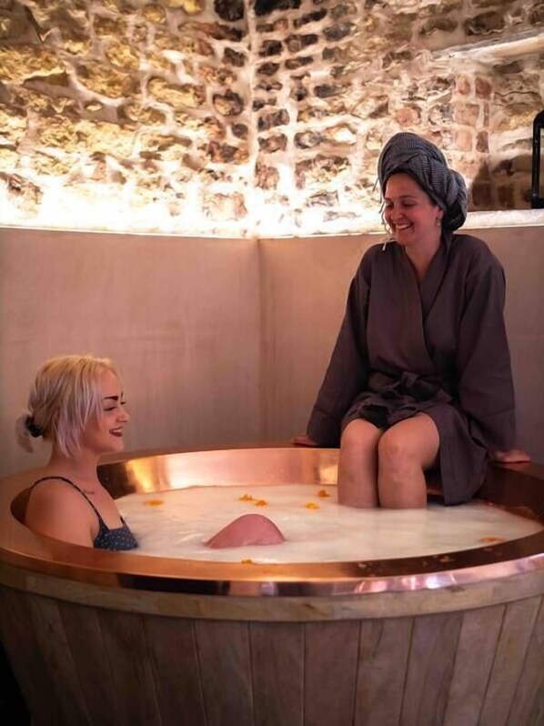 A stone-walled room with a wooden bathtub filled with water and bath products.