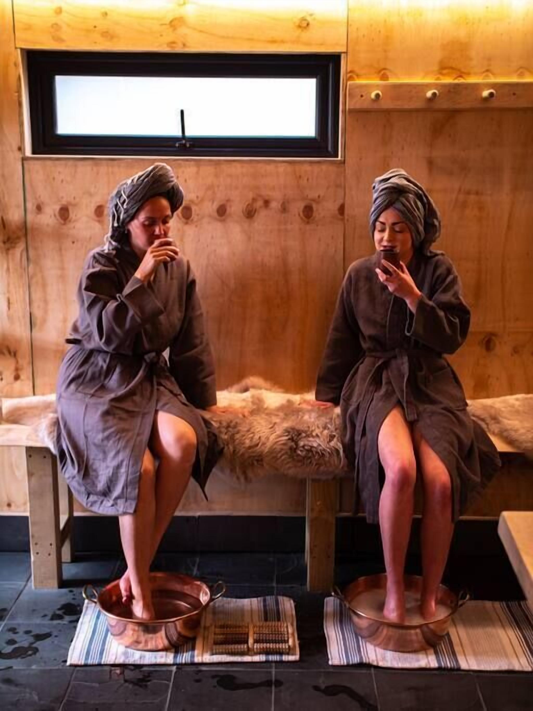 Two people sitting on a bench in a wooden room with a window, using copper basins and a striped mat on the floor.