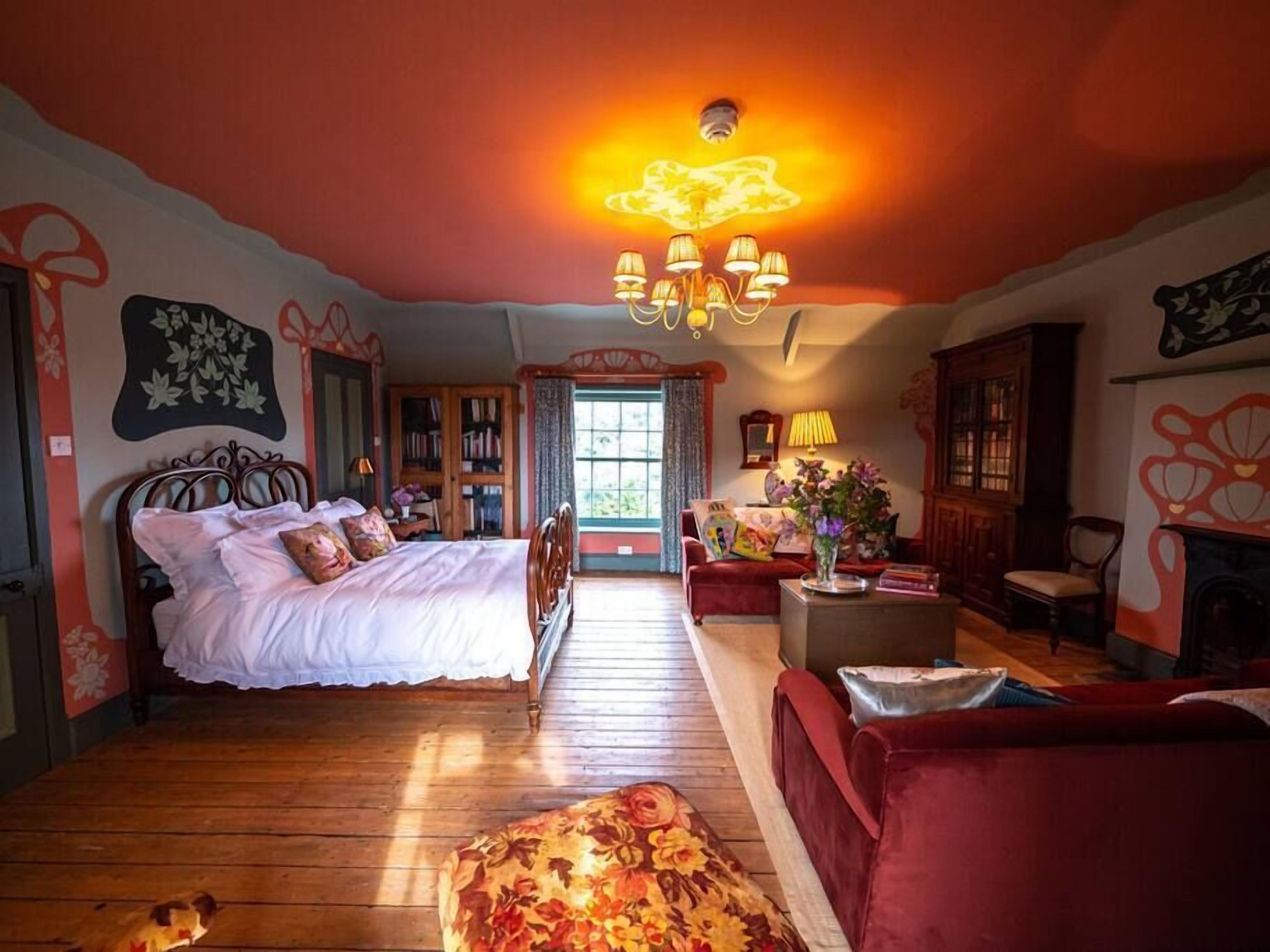 Una camera da letto con pavimento in legno, un letto con biancheria bianca, un divano rosso, un tavolino con un vaso e un lampadario.