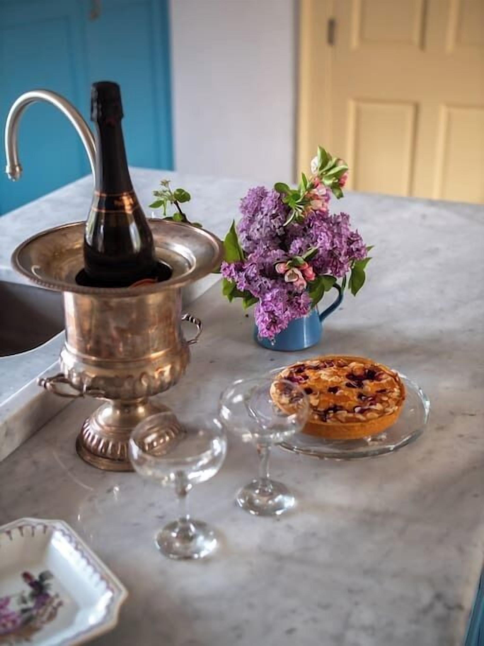 A kitchen counter with a coffee pot, a floral arrangement, a glass plate with a pastry, and wine glasses.