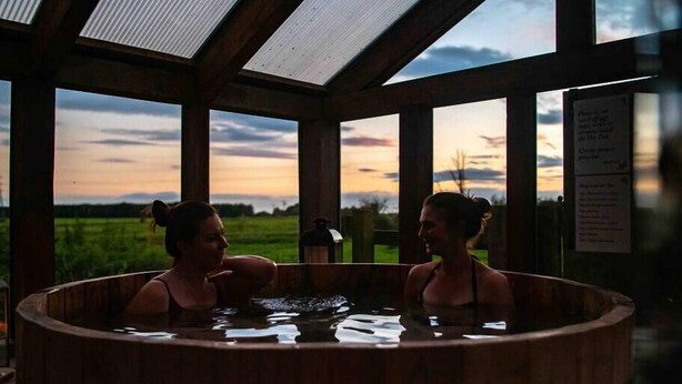 Two people in a wooden hot tub with a view of a sunset and a grassy field. - Library Suite: Sleeps 4 (Scunthorpe)