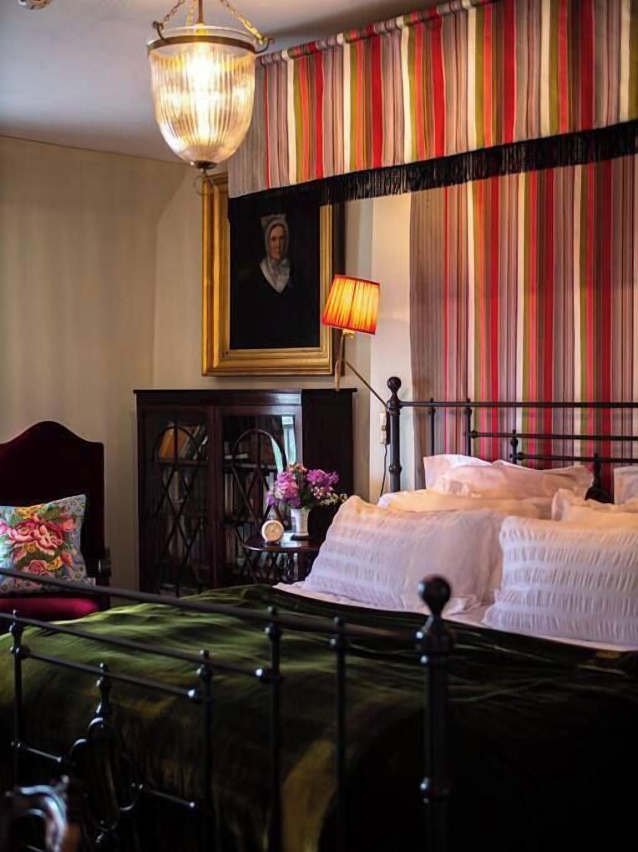A bedroom with a bed, a nightstand, a lamp, a painting, and a window with curtains.