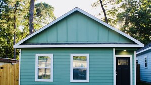 A small, single-story house with a green exterior and white trim. It features a black front door with a glass panel, two windows with white frames, and a small porch area. - The Beaver Den 30 day stays King bed Lake Conroe (Montgomery)