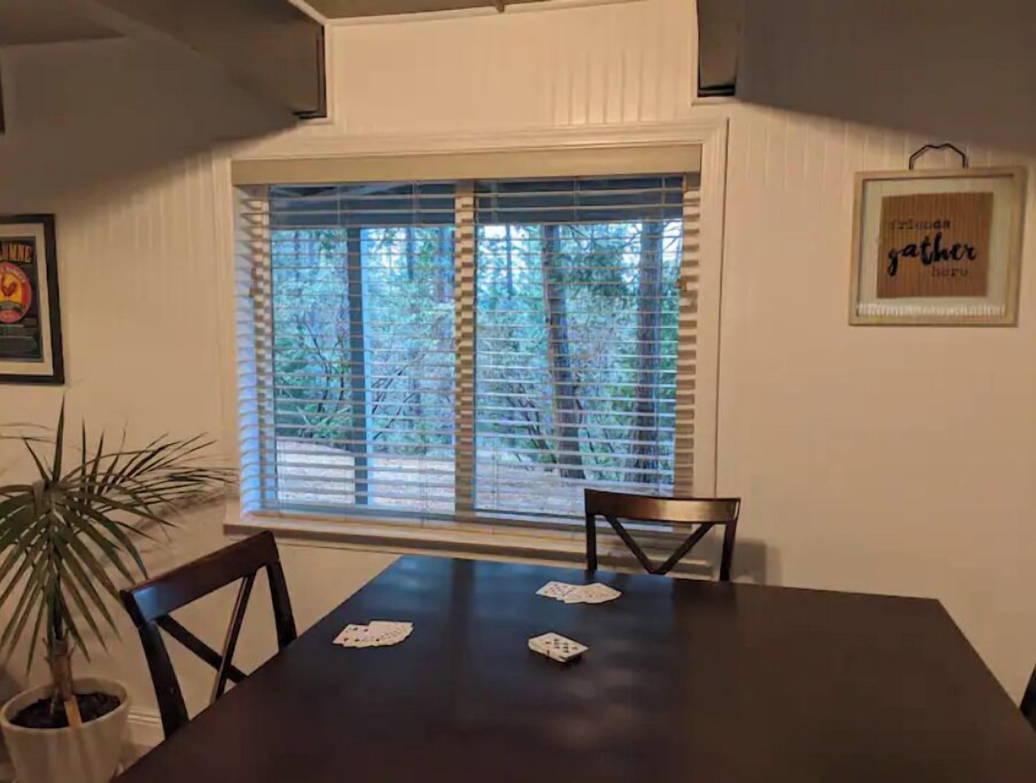 A dining area with a table set for two, a potted plant, and a window with blinds.