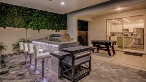 A modern outdoor kitchen area with a foosball table, bar stools, and a grill. - Breezin': Pool, Spa, Fire Pit, Foosball and more! (Indio)