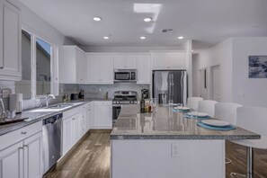 A modern kitchen with white cabinetry, a central island with blue placemats, stainless steel appliances, and a large window. - Breezin': Pool, Spa, Fire Pit, Foosball and more! (Indio)