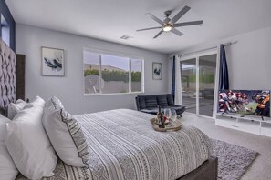 A bedroom with a bed, a ceiling fan, a large window, a TV, and a sliding glass door leading to an outdoor area. - Breezin': Pool, Spa, Fire Pit, Foosball and more! (Indio)