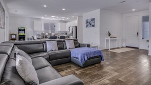 A modern living room with a sectional sofa, a dining area, and a kitchen in the background. - Breezin': Pool, Spa, Fire Pit, Foosball and more! (Indio)