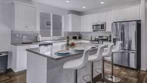 A modern kitchen with white cabinets, a stainless steel refrigerator, a built-in microwave, and a bar area with white bar stools. - Breezin': Pool, Spa, Fire Pit, Foosball and more! (Indio)