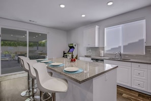 A modern kitchen with a bar area, white cabinetry, and a view through the glass door. - Breezin': Pool, Spa, Fire Pit, Foosball and more! (Indio)