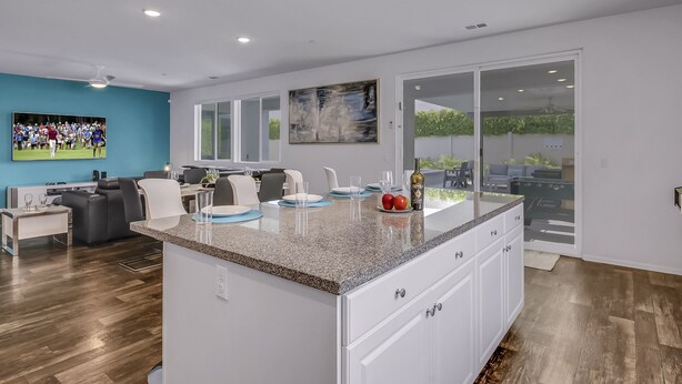 A modern kitchen with a granite countertop, white cabinets, and a large island. There is a dining area with a gray sofa and a TV mounted on the wall. - Breezin': Pool, Spa, Fire Pit, Foosball and more! (Indio)