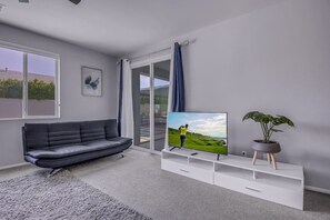 A modern living room with a grey sofa, a flat-screen TV, a potted plant, and a sliding glass door leading to an outdoor area. - Breezin': Pool, Spa, Fire Pit, Foosball and more! (Indio)